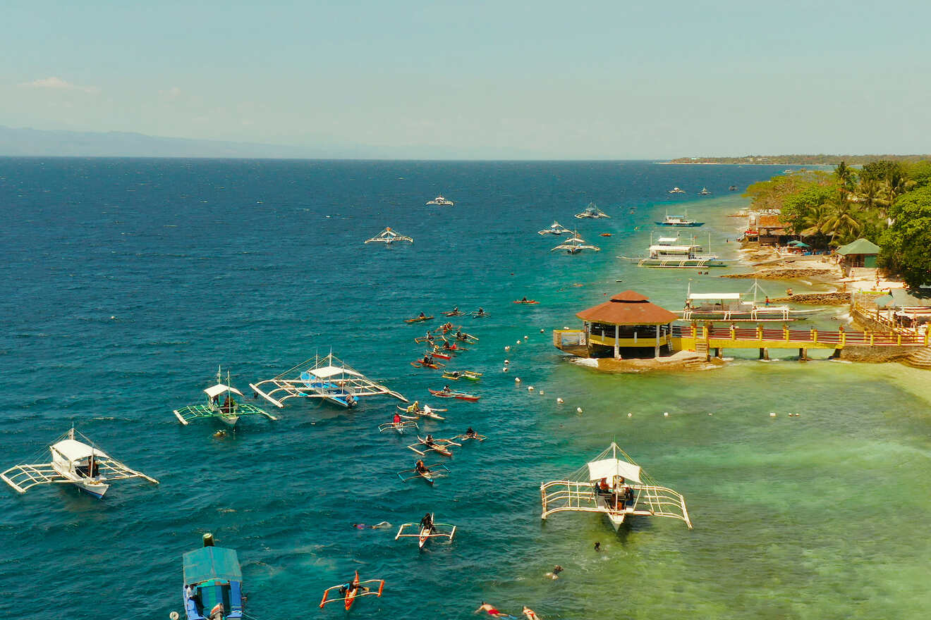 High aerial shot of a vibrant coastal area with boats in crystal clear blue waters, numerous tourists enjoying water activities around a small pier and huts.